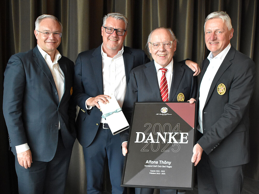 Vier Männer in Anzügen stehen eng beieinander und lächeln. Einer hält ein Bild mit einem Text, der eine Leistung feiert. Der Hintergrund ist ein dunkler, eleganter Vorhang.