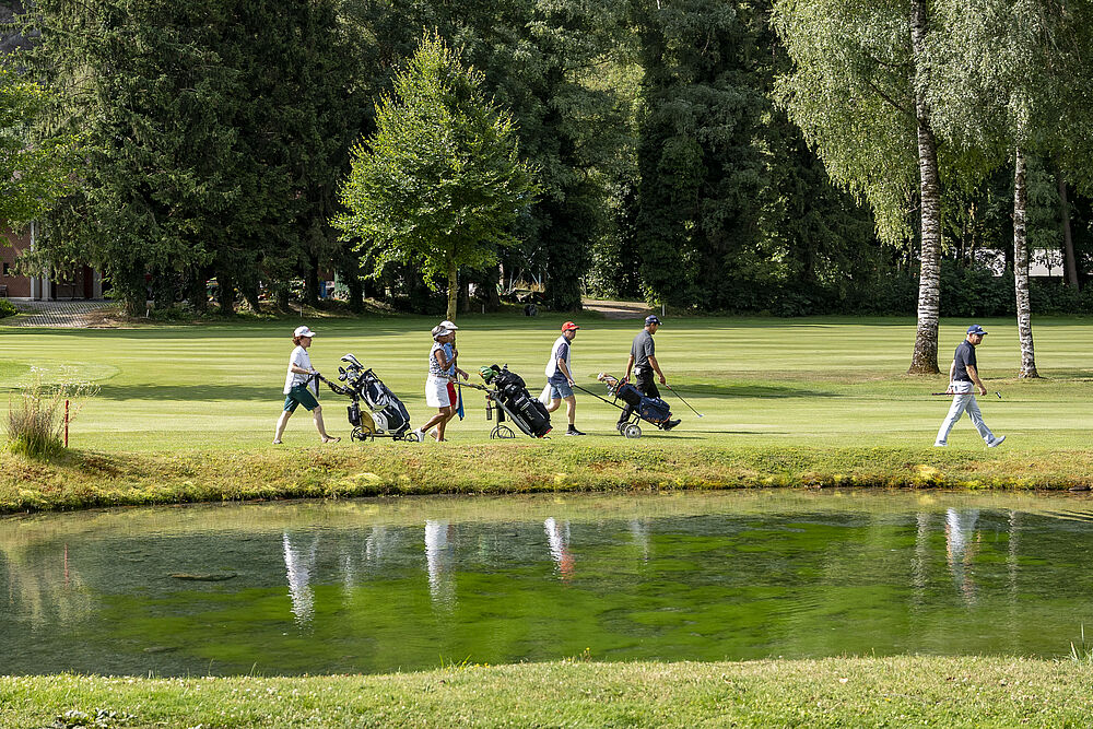 Eine Gruppe von fünf Golfspielern geht über einen üppig grünen Golfplatz, jeder zieht eine Golftasche. Im Vordergrund ist ein spiegelnder Teich zu sehen, im Hintergrund stehen hohe Bäume.