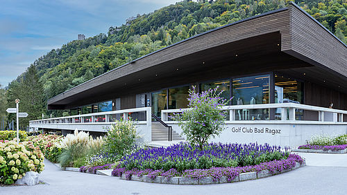 Das Clubhaus des Golf Club Bad Ragaz. Ein modernes Gebäude mit Flachdach, umgeben von Gärten an einem Berghang. Blumen im Vordergrund, Wald im Hintergrund, hell und heiter.