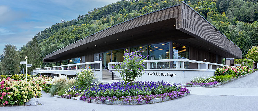 The clubhouse of the Bad Ragaz Golf Club. A modern building with a flat roof, surrounded by gardens on a mountainside. Flowers in the foreground, forest in the background, bright and cheerful.