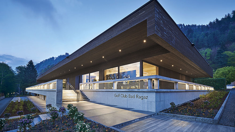 Modern building with a sharp, angular flat roof against a dusky sky and mountain backdrop. The soft lighting creates a warm, inviting ambience.