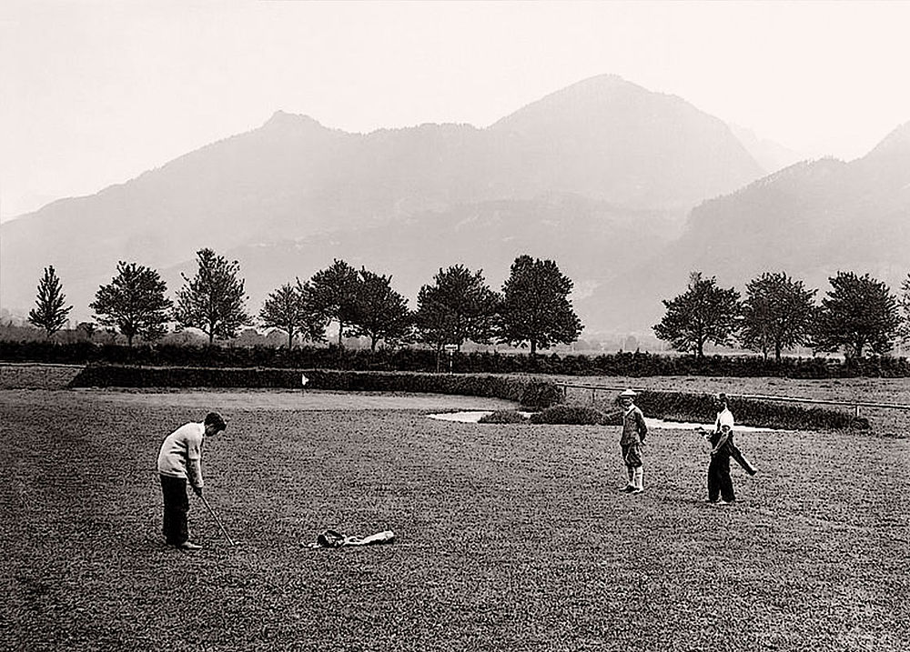 Schwarz-Weiß-Foto von drei Menschen, die auf einem Feld Golf spielen, mit einer Reihe von Bäumen und nebligen Bergen im Hintergrund, die eine heitere ländliche Szene vermitteln.
