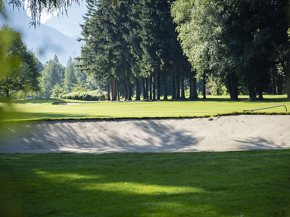 Eine sonnige Landschaft mit einem Sandbunker im Vordergrund auf einem Golfplatz, umgeben von saftig grünem Gras und hohen Bäumen vor einem strahlend blauen Himmel mit vereinzelten Wolken.