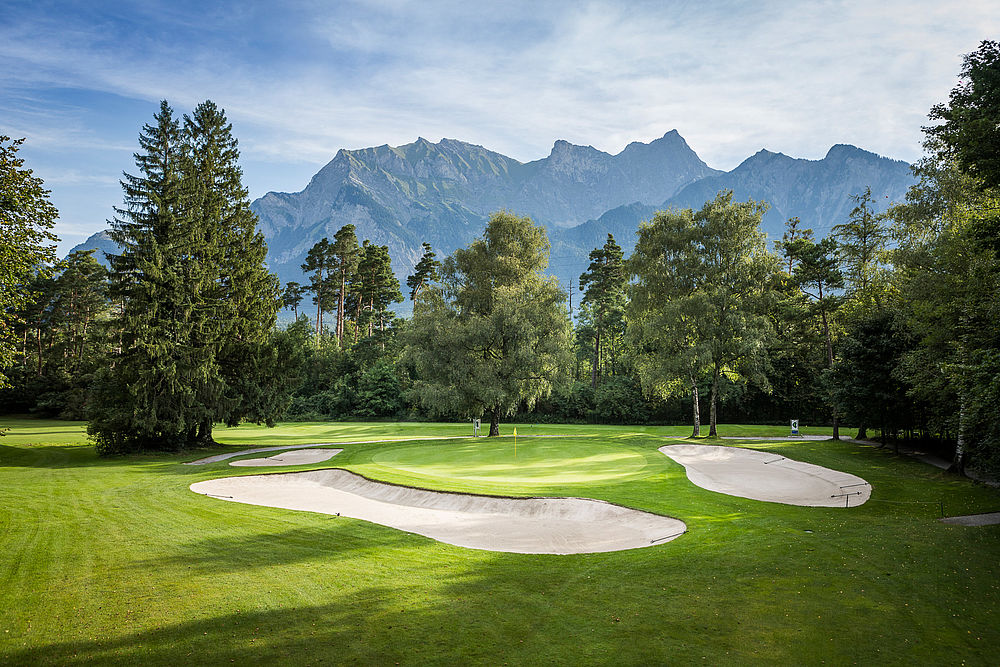 Golfplatz mit Sandfängen, grünem Fairway und hohen Bäumen. Berge im Hintergrund unter einem klaren blauen Himmel. Ruhige, malerische Landschaft.