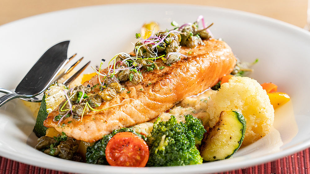 Gegrillter Lachs mit Kräutern auf einem Bett aus Brokkoli, Zucchini, Kirschtomaten und Kartoffelpüree. Gabel und Messer am Rand des Tellers.