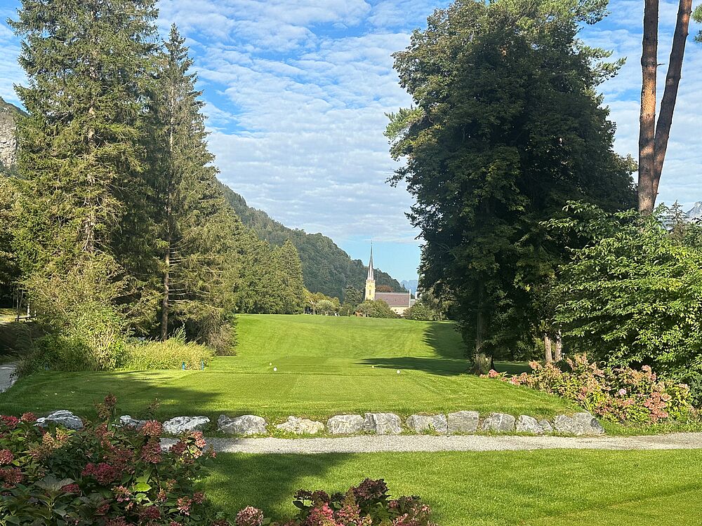 Mit Blick auf den Kirchturm von Bad Ragaz steht man auf dem Abschlag. Grüne wiese im Vordergrund. Links und rechts umringt von Bäumen. 