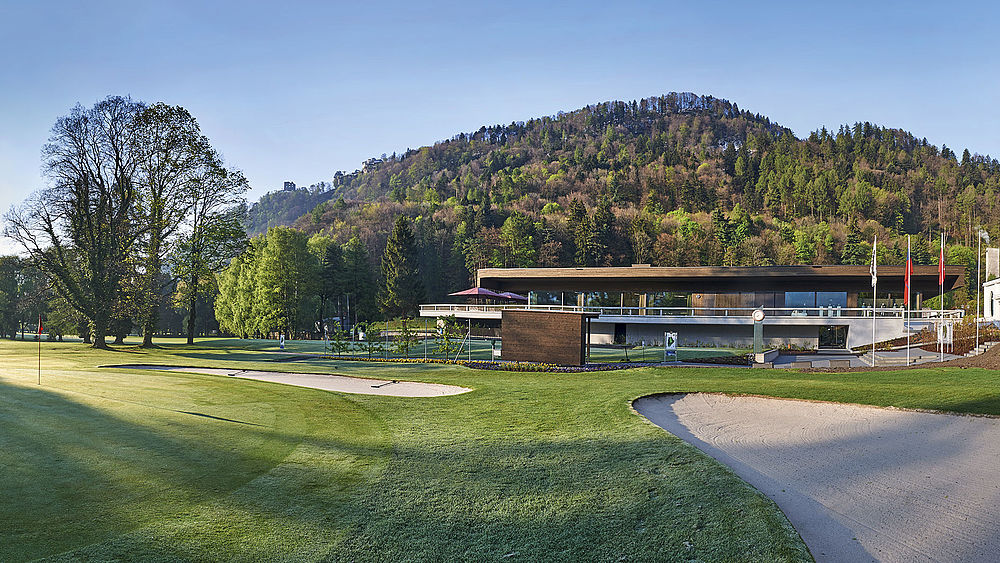 The modern clubhouse with large windows and terrace is nestled in a green landscape directly behind tee 1. The mountains and the blue sky form a serene backdrop that radiates tranquillity.