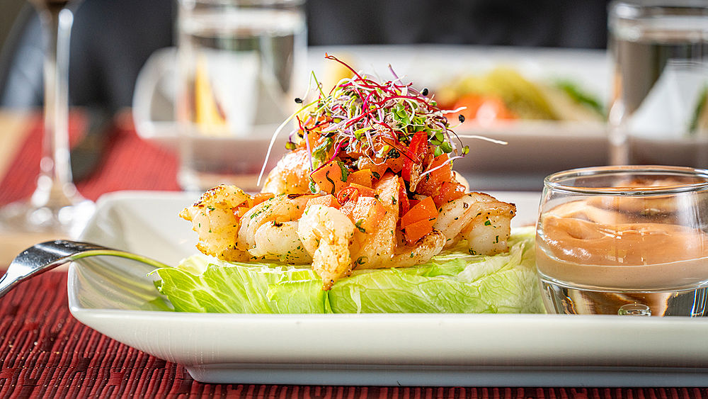 Garnelencocktail auf Kopfsalat, garniert mit Sprossen und Sesam, serviert mit einer Dip-Sauce. Elegantes Restaurant im Hintergrund.