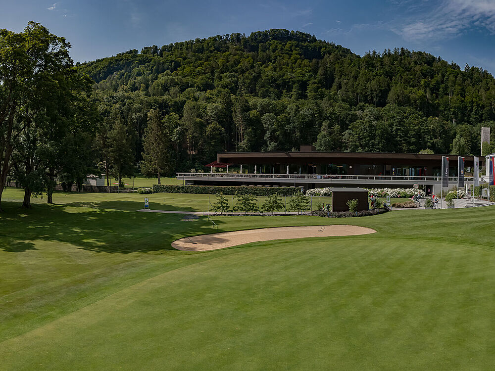 Panoramablick auf einen Golfplatz mit Sandbunkern, umgeben von Bäumen und Hügeln. In der Ferne sind ein modernes Clubhaus und Menschen zu sehen.
