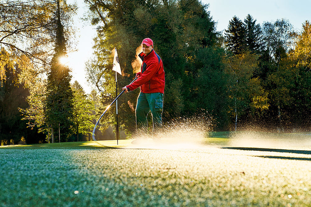 Eine Person harkt bei Sonnenaufgang einen Bunker auf einem Golfplatz und wirft dabei lange Schatten. Die leuchtend rote Jacke kontrastiert mit dem grünen Gras und schafft eine friedliche Szene bei Sonnenaufgang.