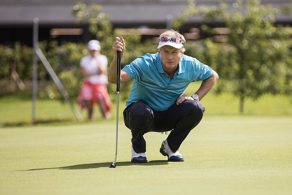 Golfer in blauem Hemd hockt, konzentriert auf einen Putt auf einem sonnigen Grün. Der Hintergrund zeigt eine verschwommene Figur und Bäume, die Konzentration und Ruhe vermitteln.